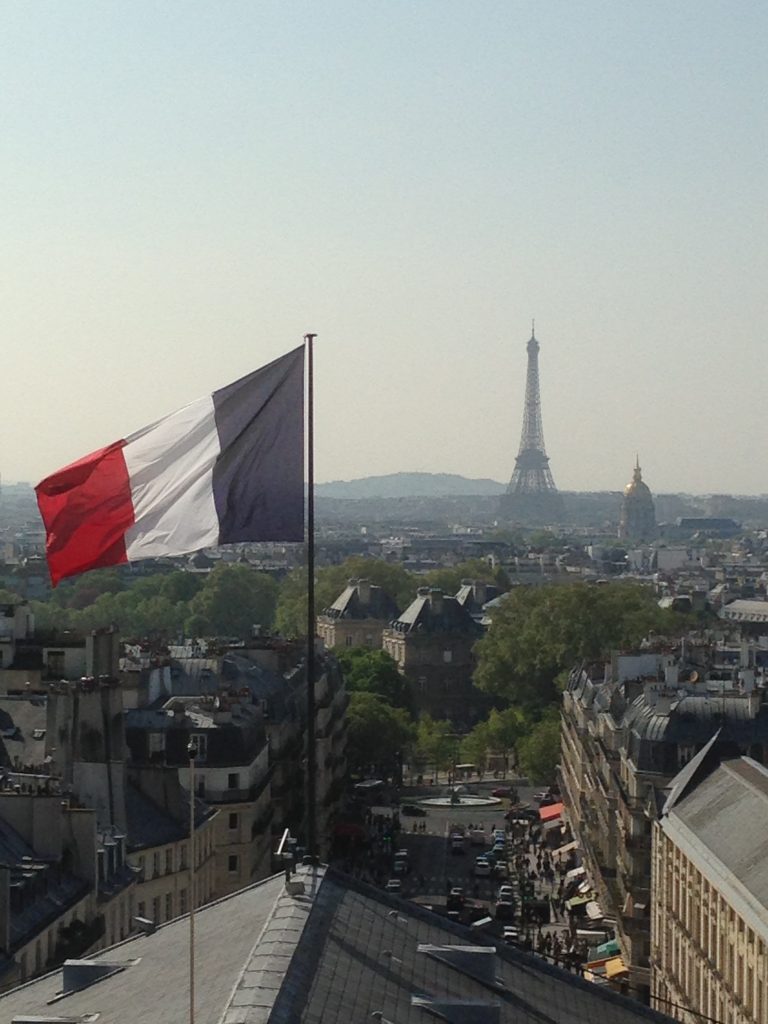 A photo of the landscape of Paris. The Eiffel Tower is in the distance and a French flag is waving in the foreground.