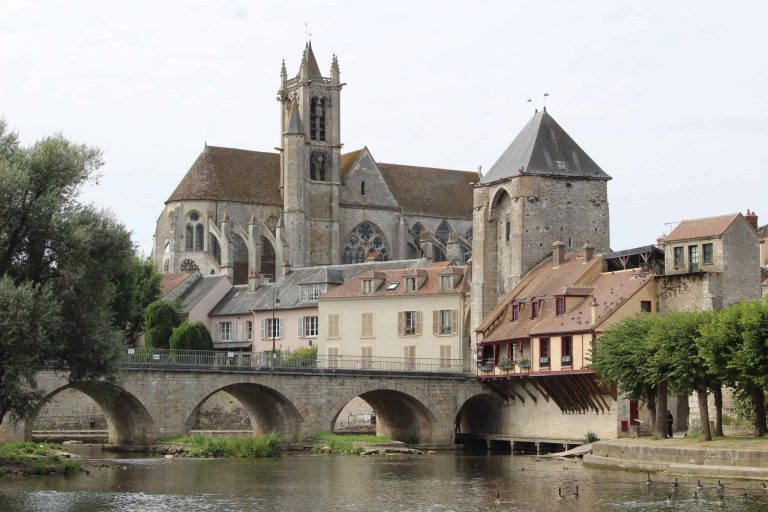 A photo of the town of Moret-sur-Loing in the suburbs of Paris. It shows a bridge going over a river, with a medieval church in the background.