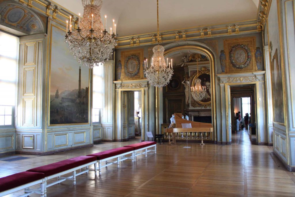 A photo of the Salle des Fetes, looking down towards the harpsichord in the musician's gallery at the end. The floors are wooden, the walls are covered in paintings, and three large chandeliers hang from the ceiling.