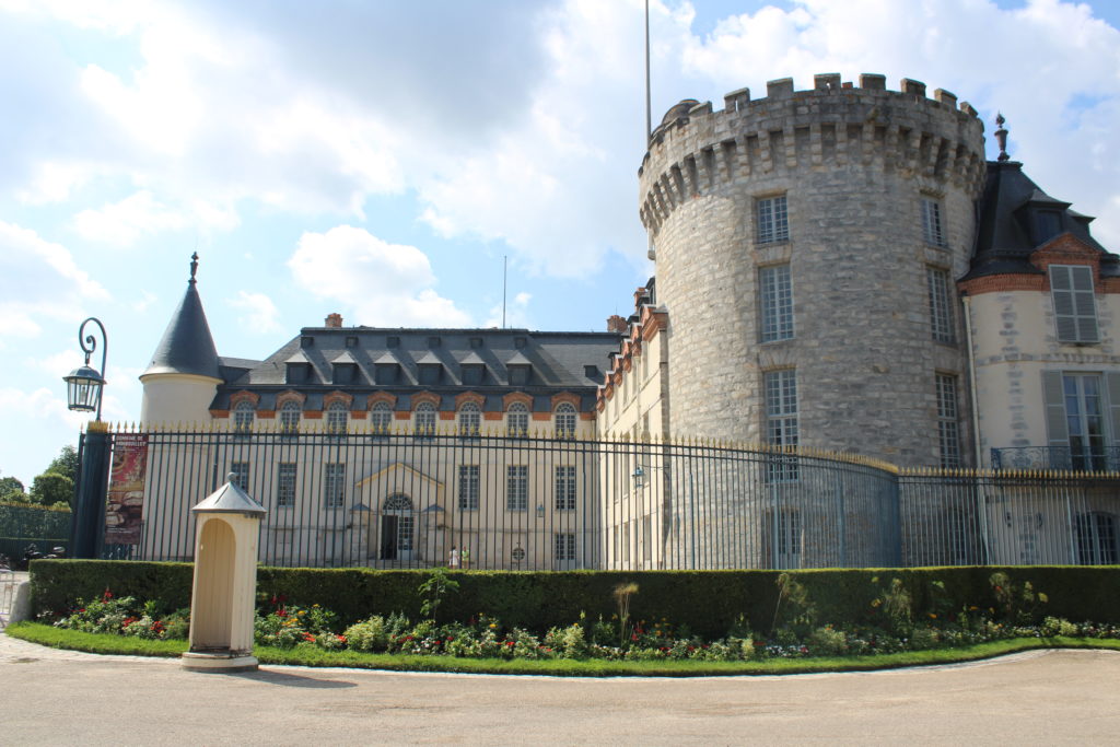 A photo of the exterior facade of the Château de Rambouillet, looking at the front courtyard surrounded by a fence. 