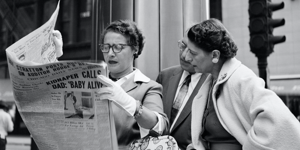 A black and white photograph of two women looking at a newspaper.