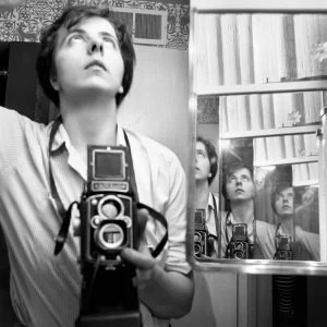 A black and white photograph from the Vivian Maier exhibition at the Musée du Luxembourg. The photo is a self portrait of Maier. She is holding a camera to her chest, facing a mirror, and looking up.