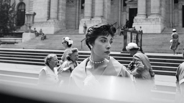 A black and white photo of an elegantly dressed woman standing in front of the New York City library. She is looking away from the camera, over her left shoulder down the street.