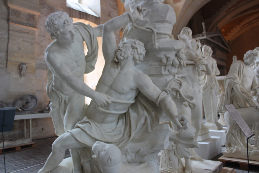 A photo of a sculpture in the Galerie des Sculptures et des Moulages. The sculpture is of one man tying another man to the rocky cliff behind him.