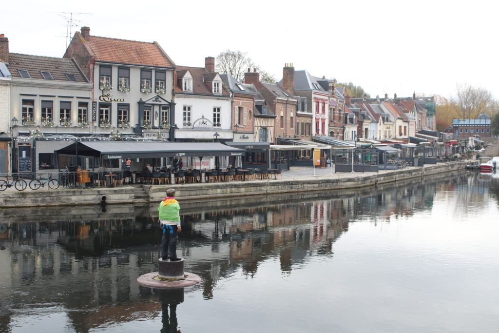 A photo of Saint-Leu street in Amiens. There are several restaurants and shops along the water, and a statue of a man on a pillon in the middle of the water.