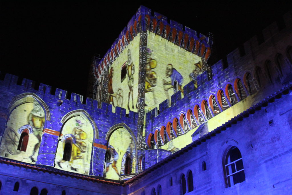A photo of the Palais des Papes in Avignon at night, illuminated by digital projects on a corner tower.