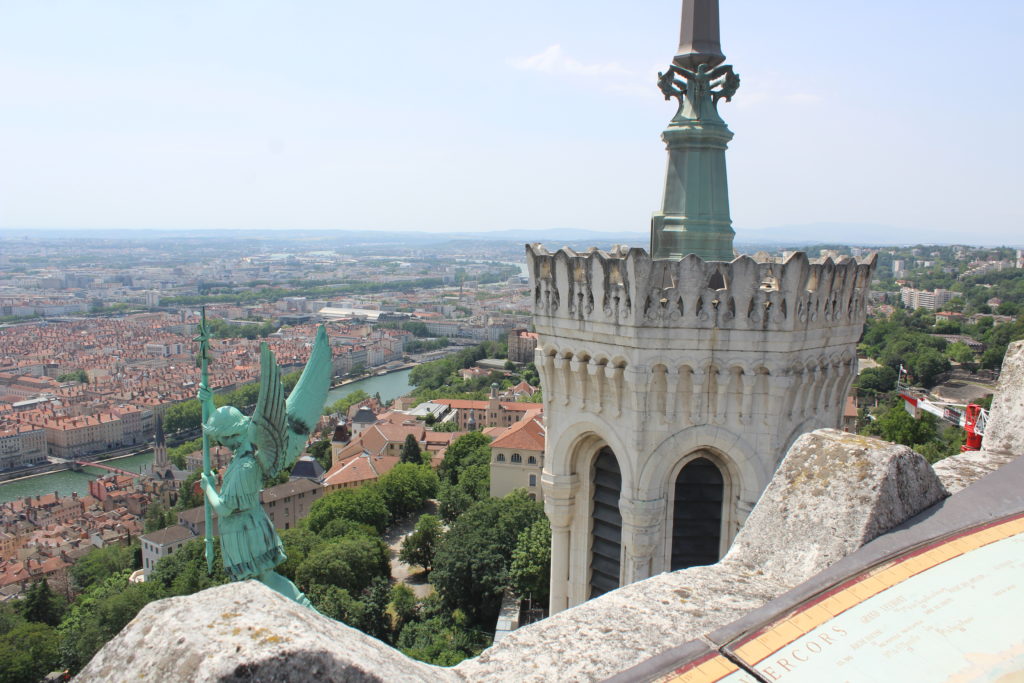 A photo of Lyon in the distance, taken from the roof of the Basilica of Notre-Dame de Fourvière. The basilica tower and a green statue are in the foreground.