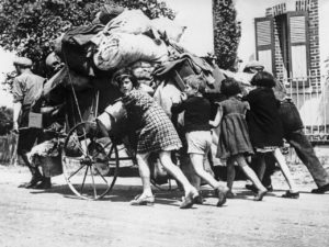 A black and white photo from the Exodus of Paris. Several children are pushing a cart laden down with their belongings.