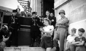 A photo of Simone Segouin, a French Resistance fighter, standing on some steps, holding a machine gun, while several men and boys watch her.