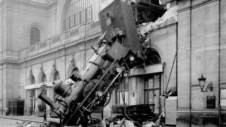 A black and white photo of a train locomotive that has burst from the wall to land on the street one floor below.