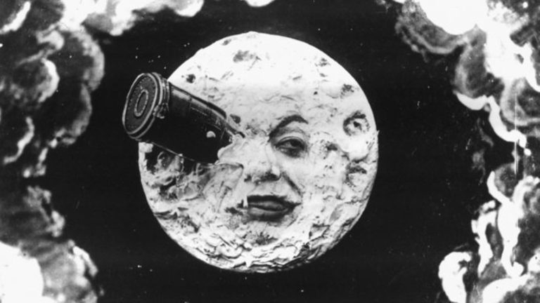 A black and white image from A Trip to the Moon by Georges Méliès. A rocket ship is sticking out of the eye of the Man on the Moon.