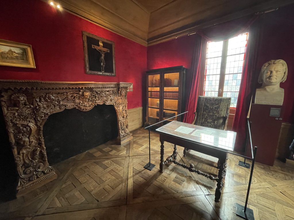 A photo of the writing room in the Maison de Balzac. There is a desk with a chair, a fireplace, bookshelves, and a bust of Balzac on the walls.