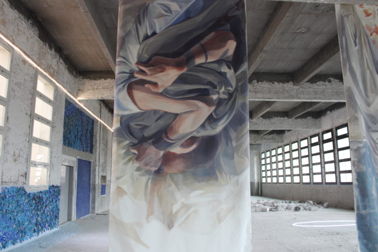 A photo of a floor to ceiling painting in an abandoned building. The paint shows two arms hugging a blanket.
