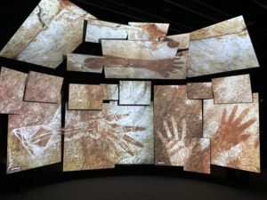 A photo of an exhibition at the Musée de l'Homme. It shows a bank of screens, all arranged in an arch, with images of prehistoric hands displayed on them.
