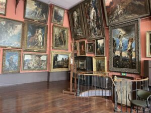 A photo of the interior of the Musee National Gustave Moreau. It shows a corner of the studio space. The walls are red and every inch is covered in artwork.