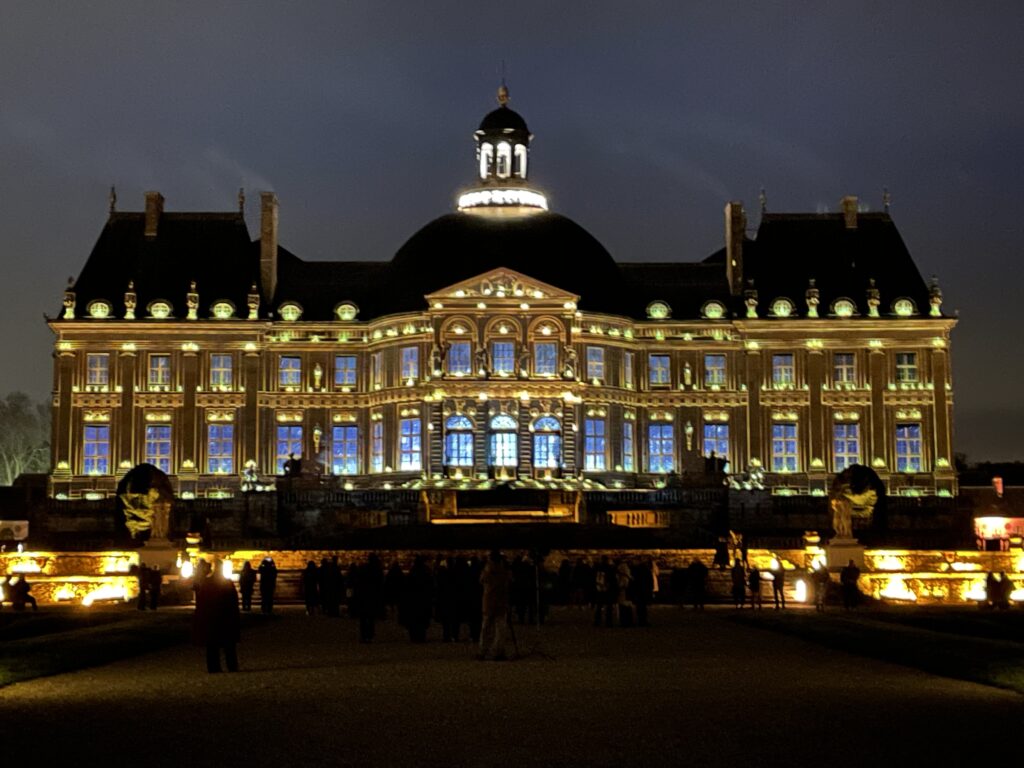 A photo of the exterior facade of Vaux-Le-Vicomte. The building is illuminated at night with projections and lights.