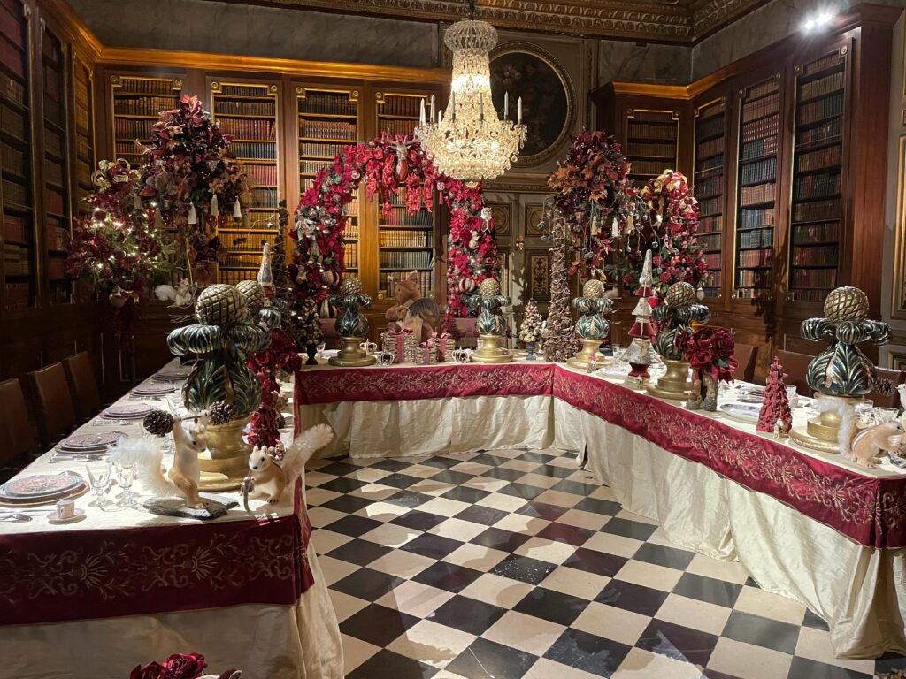 A photo of one of the Christmas displays in the library. A large, U-shaped table is decorated with place settings, wreaths, and holiday ornaments.