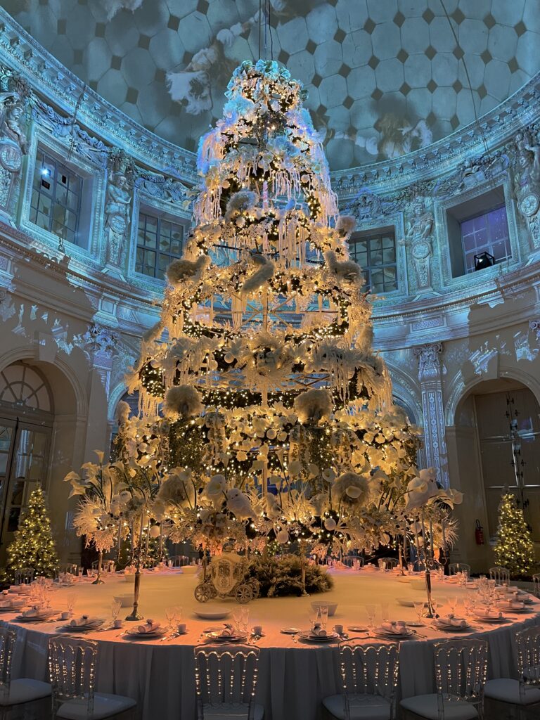 A photo of a magnificent tree in the main foyer of the chateau. It is sitting on a large round table that has place settings all along it. The walls behind are bathed in blue light and projections.
