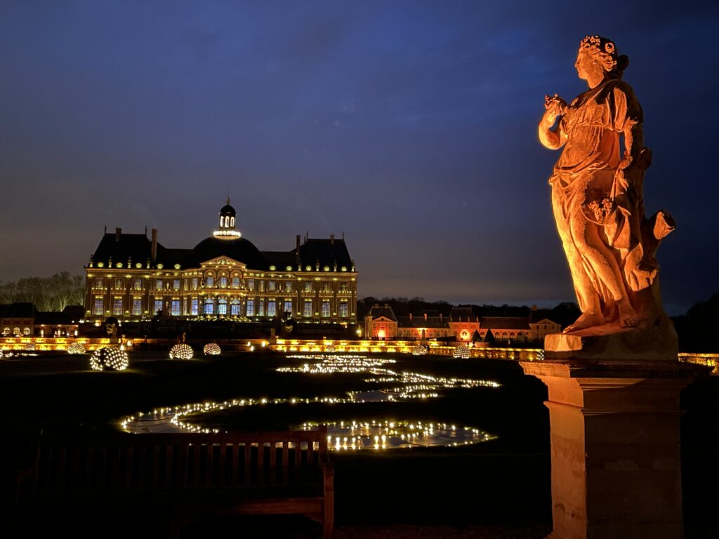 A photo of the back garden of the chateau. An illuminated statue of a goddess is in the forefront, and the ponds and gardens are illuminated with lights. The illuminated chateau is in the background.